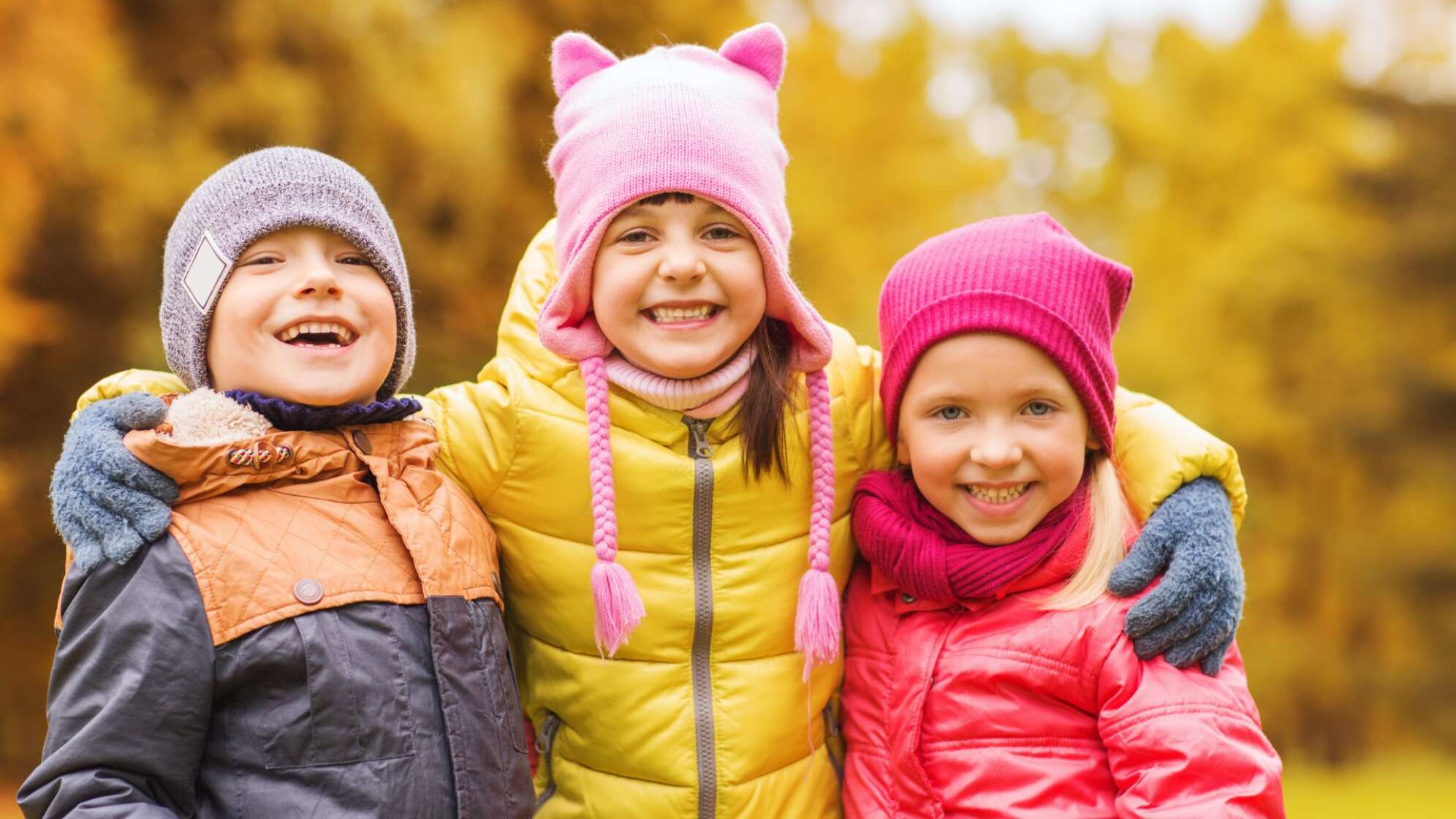 Drei glücklich strahlende Kinder mit Jacken und Mützen vor einem herbstlichen Park