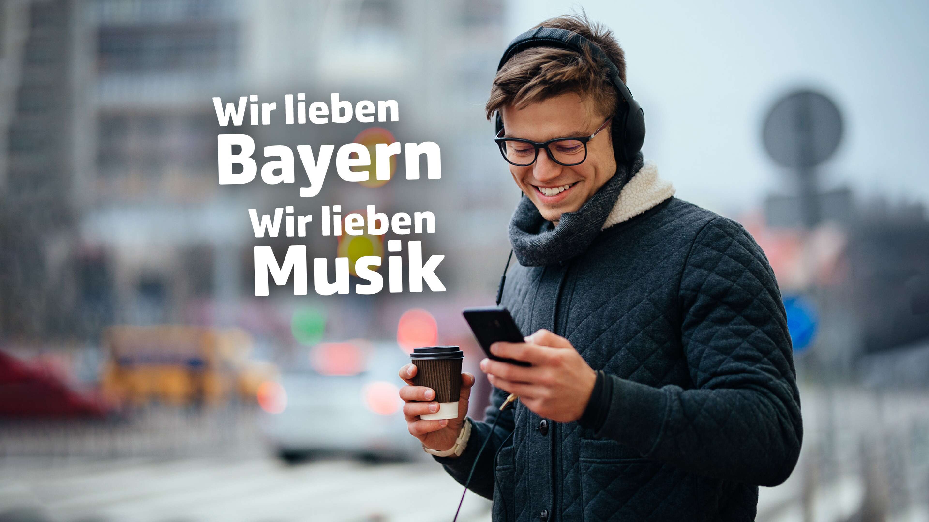 Mann mit Winterklamotten steht draußen und blickt lächelnd auf sein Smartphone, dazu der Text: Wir lieben Bayern, wir lieben Musik