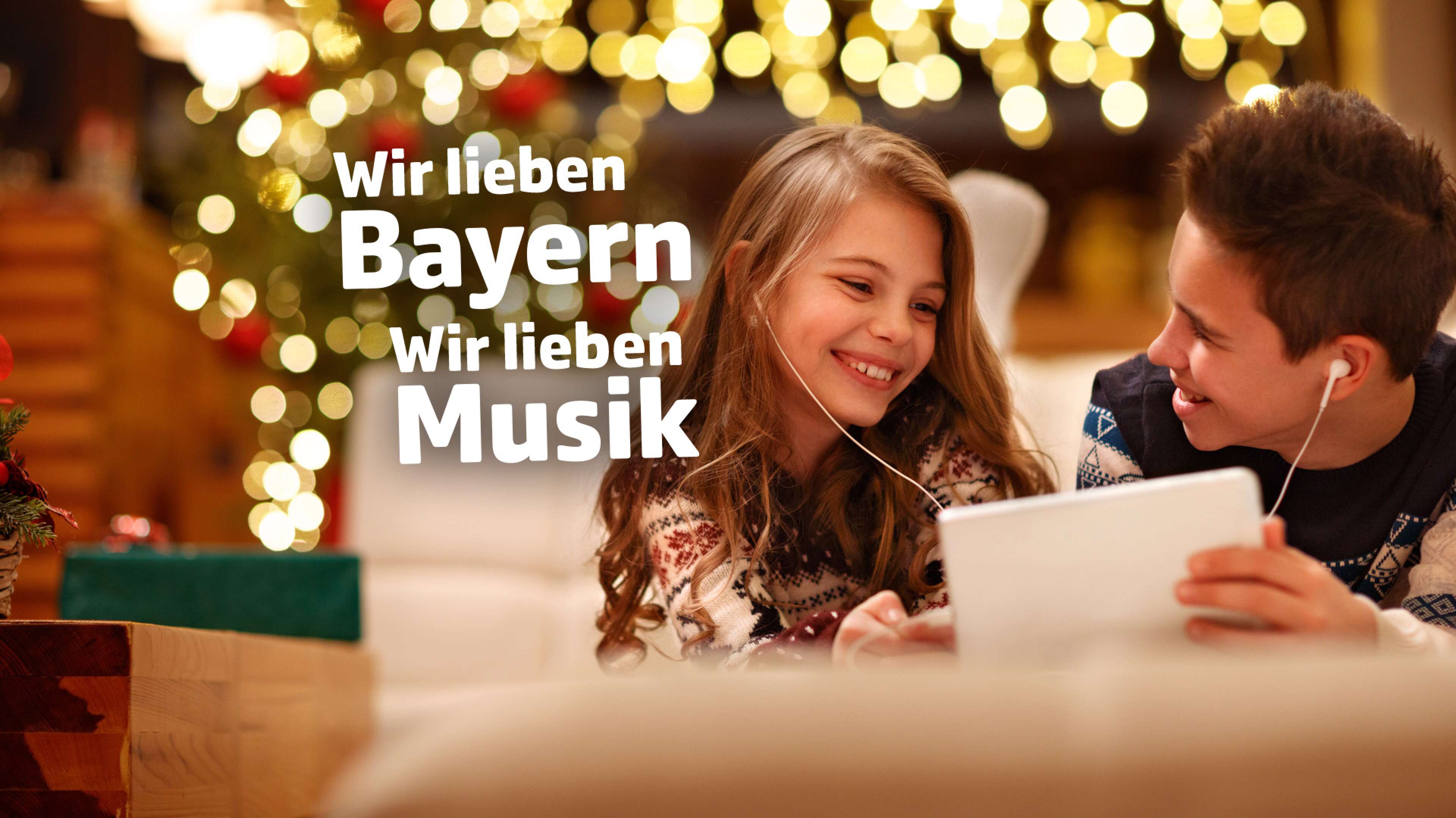 Ein Junge und ein Mädchen liegen auf der Couch und hören gemeinsam mit Kopfhörern Musik auf einem Tablet, im Hintergrund verschwommen ein Christbaum, dazu der Text: Wir lieben Bayern, wir lieben Musik
