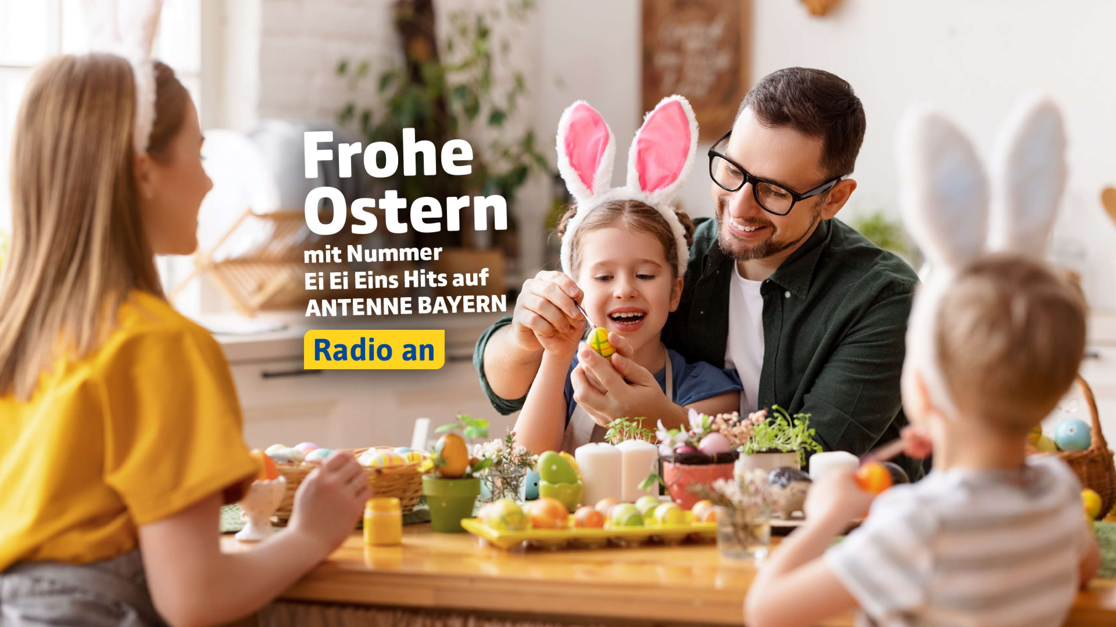 Eine glücklich lachende Familie am Oster-Tisch, ein kleines Mädchen trägt Hasenohren - dazu der Text: Frohe Ostern mit Nummer Ei Ei Eins Hits auf ANTENNE BAYERN, Radio an
