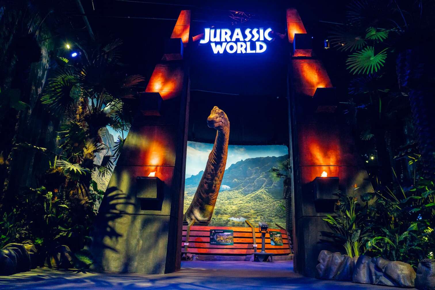 Eingangsbereich der Jurassic World Ausstellung mit beleuchtetem Jurassic World Schriftzug, tropischer Pflanzenkulisse und einem großen Dinosaurier-Modell vor einer Landschaftskulisse