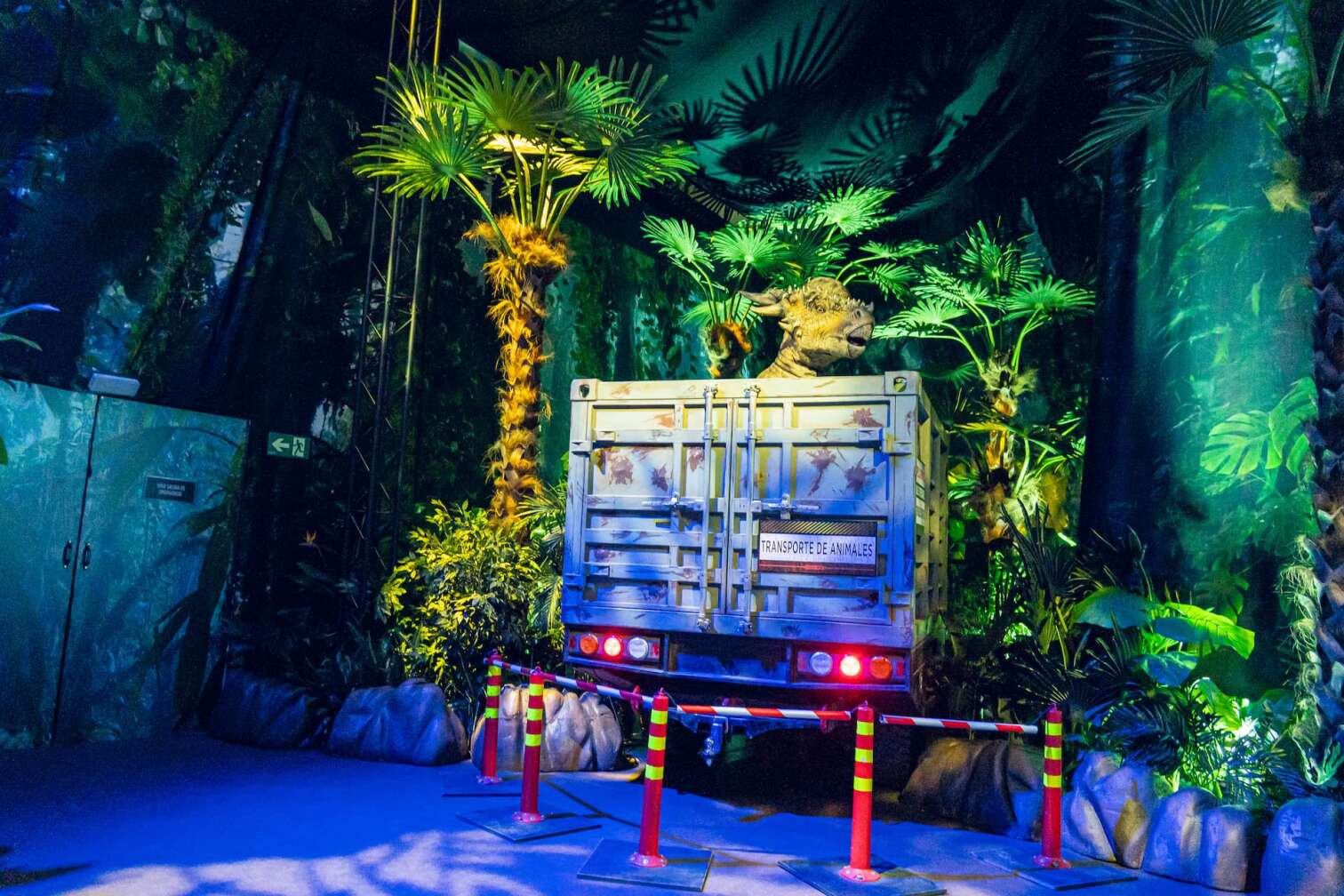 Dinosauriermodell im Transportcontainer mit der Aufschrift ‚Transporte de Animales‘, umgeben von tropischer Dschungelkulisse in der Jurassic World Ausstellung