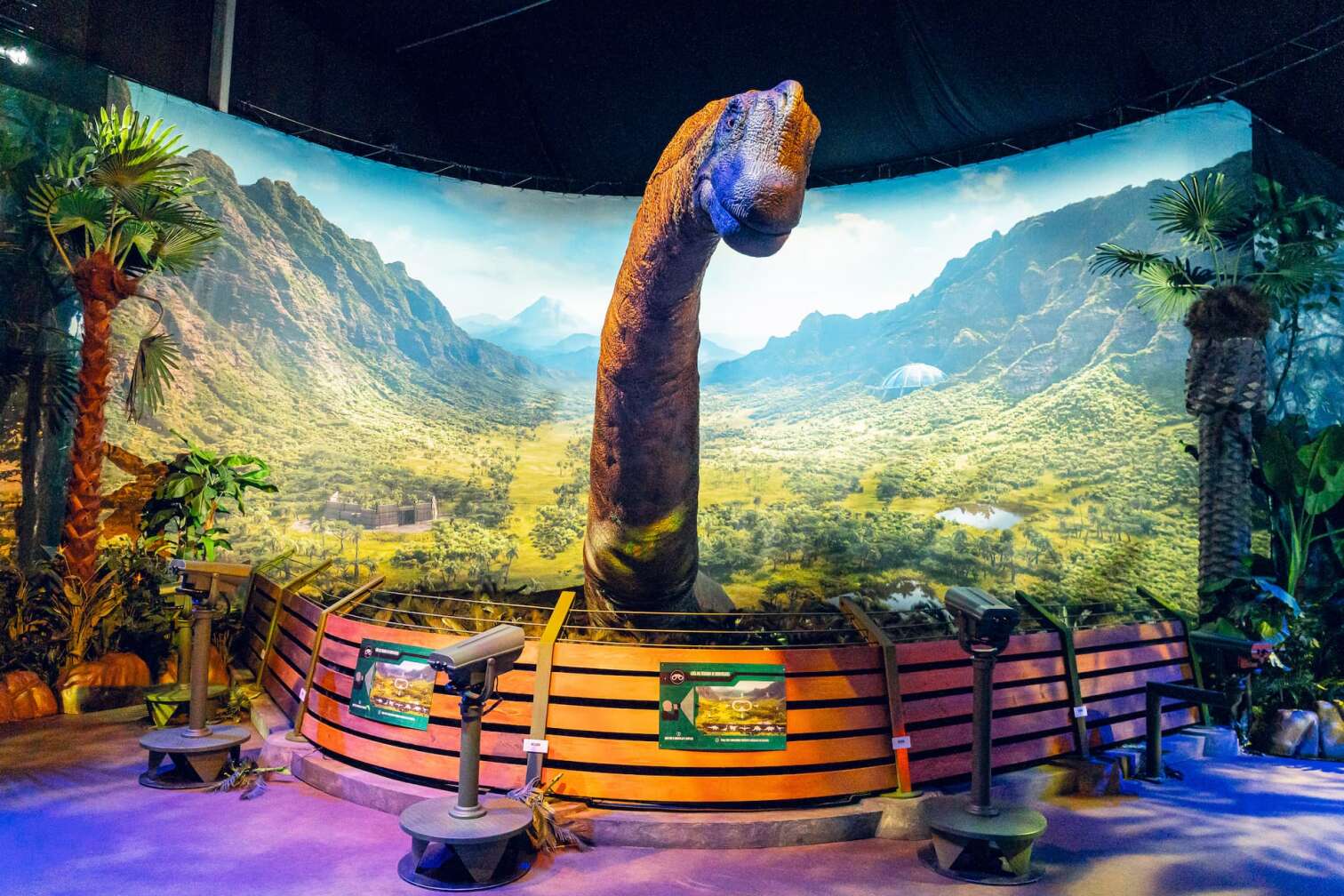 Lebensechtes Dinosauriermodell mit langem Hals vor einer tropischen Landschaftskulisse, umgeben von Ferngläsern für Besucher in der Jurassic World Ausstellung