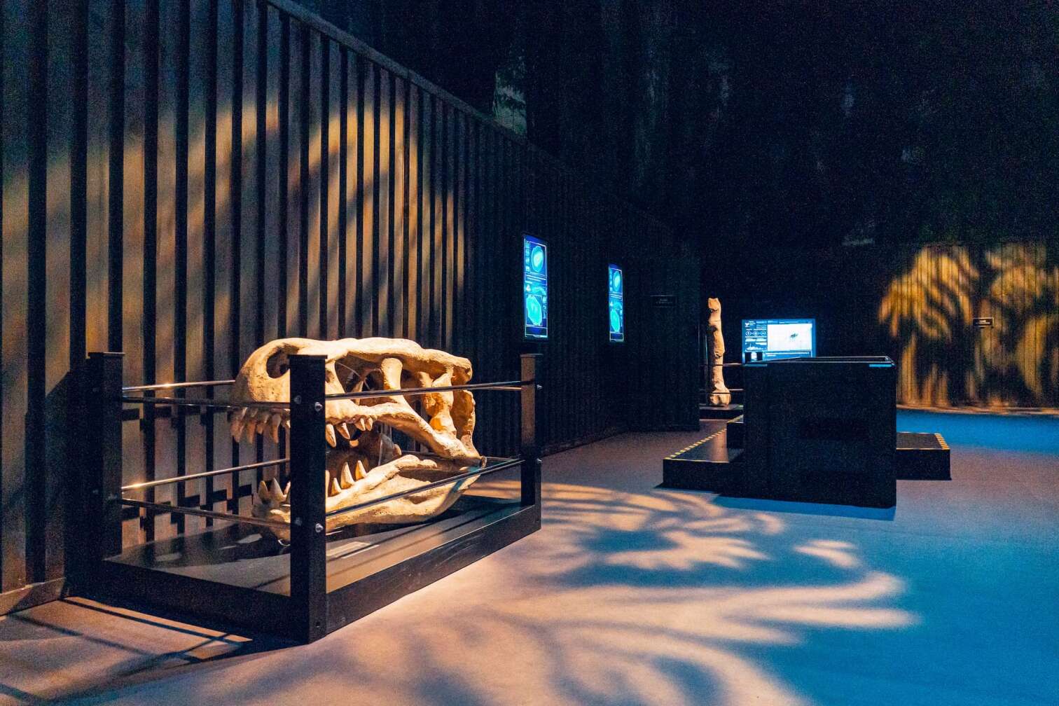 Ausstellungsraum der Jurassic World mit großem Dinosaurierschädel-Modell in einem Schutzkäfig, beleuchteten Infotafeln und moderner Präsentation