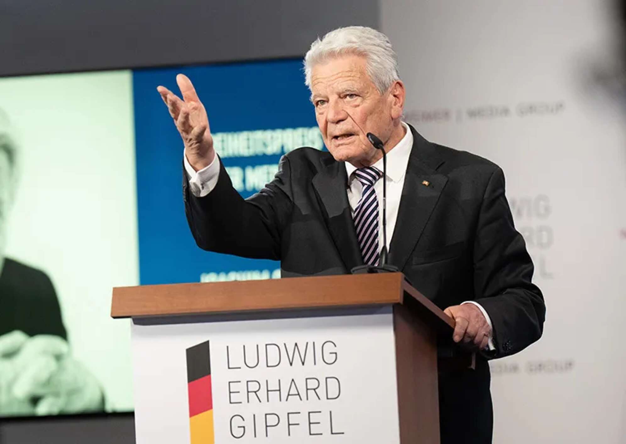 „Joachim Gauck, ehemaliger Bundespräsident, hält eine Rede am Rednerpult beim Ludwig Erhard Gipfel am Tegernsee in Bayern. Er trägt einen dunklen Anzug mit gestreifter Krawatte, gestikuliert mit der Hand und steht hinter dem Logo des Ludwig Erhard Gipfels. Im Hintergrund sind Veranstaltungsbanner und eine Leinwand sichtbar. Wirtschaftskonferenz und Politik-Event in Oberbayern.“