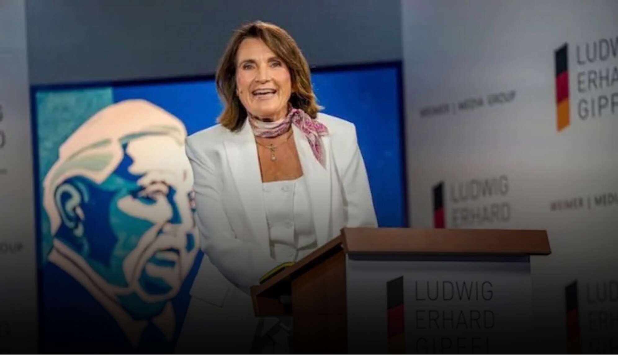 „Rednerin am Podium beim Ludwig Erhard Gipfel am Tegernsee in Bayern. Die Frau trägt ein weißes Kostüm und ein Halstuch, steht hinter einem Rednerpult mit dem Logo des Ludwig Erhard Gipfels. Im Hintergrund sind Veranstaltungsbanner und eine Illustration von Ludwig Erhard sichtbar. Wirtschaftskonferenz und Politik-Event in Oberbayern.“