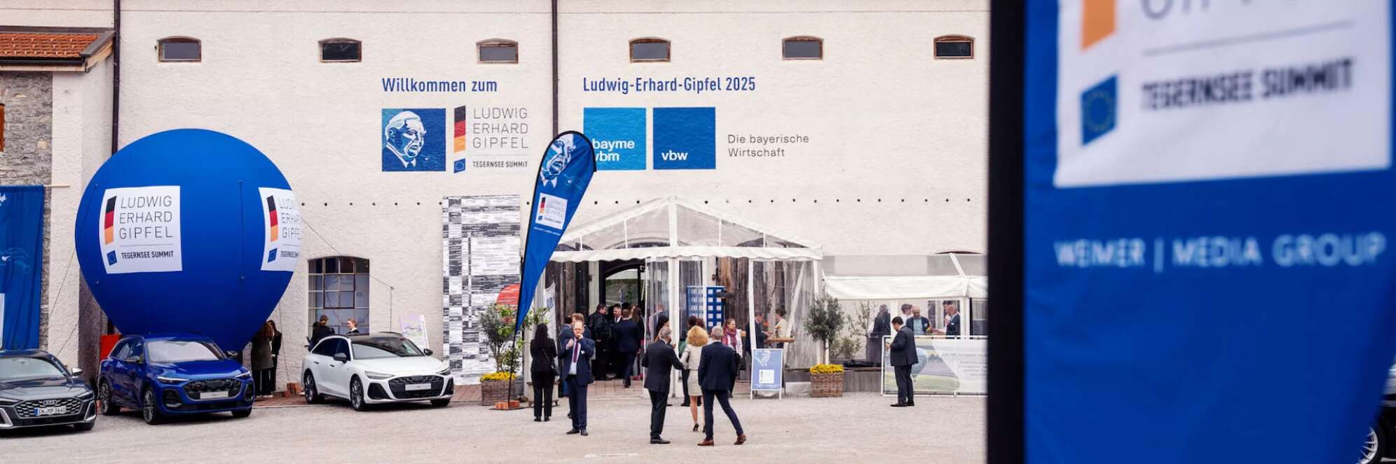 Eingangsbereich des Ludwig Erhard Gipfel 2025 am Tegernsee in Bayern. Vor dem historischen Gebäude mit Aufschrift ‚Willkommen zum Ludwig-Erhard-Gipfel 2025‘ stehen moderne Autos, ein großer blauer Ballon mit Logo und mehrere Besucher im Anzug. Banner und Logos von Sponsoren wie bayme vbm und vbw sind sichtbar. Wirtschaftskonferenz und Networking-Event in Oberbayern.
