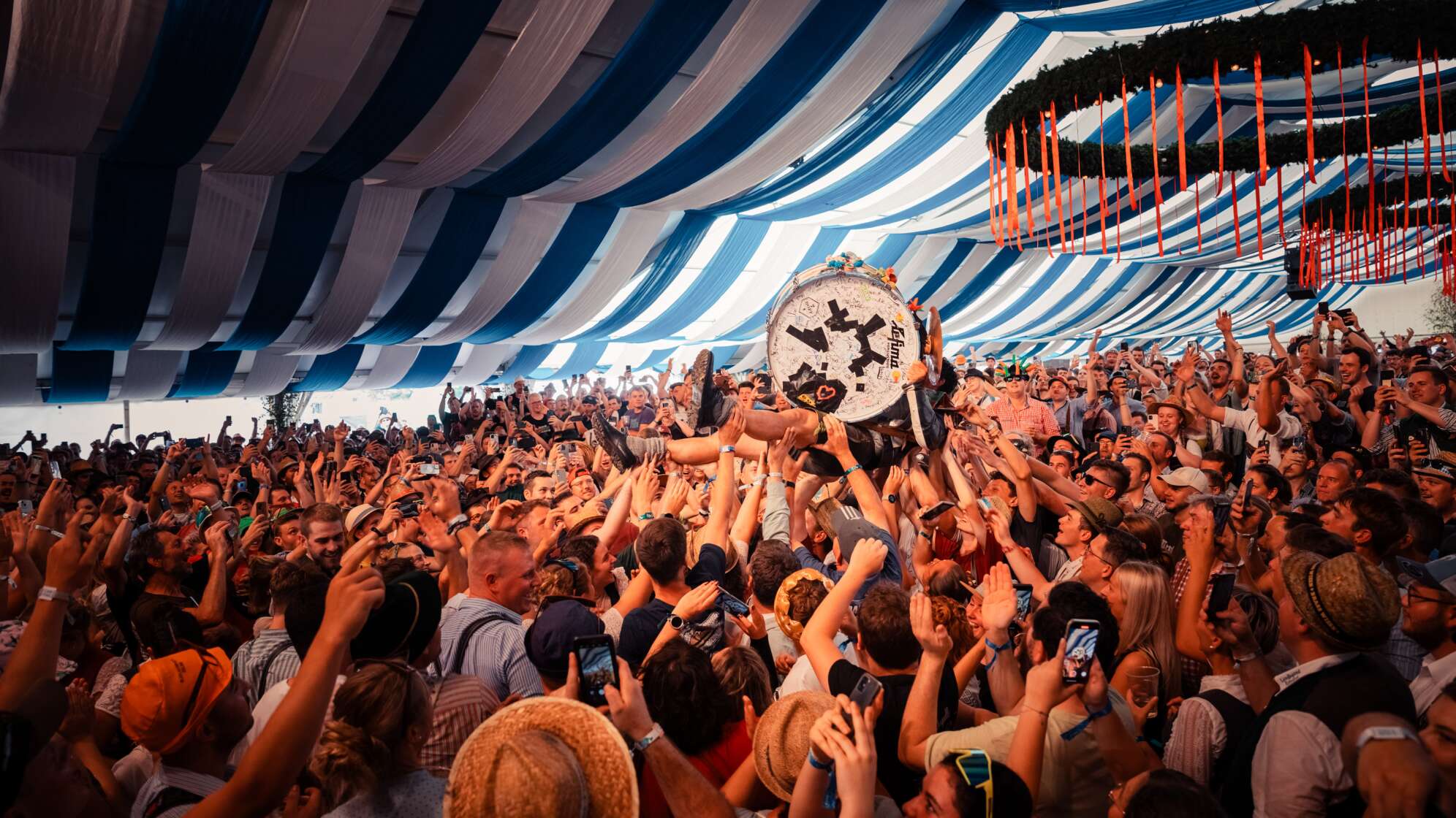 Brass Wiesn - Menschenmenge in einem blau-weiß dekorierten Festzelt bei einem Konzert oder Volksfest. Eine Person wird crowdgesurft, während viele Besucher jubeln, ihre Hände hochhalten und mit Smartphones filmen.