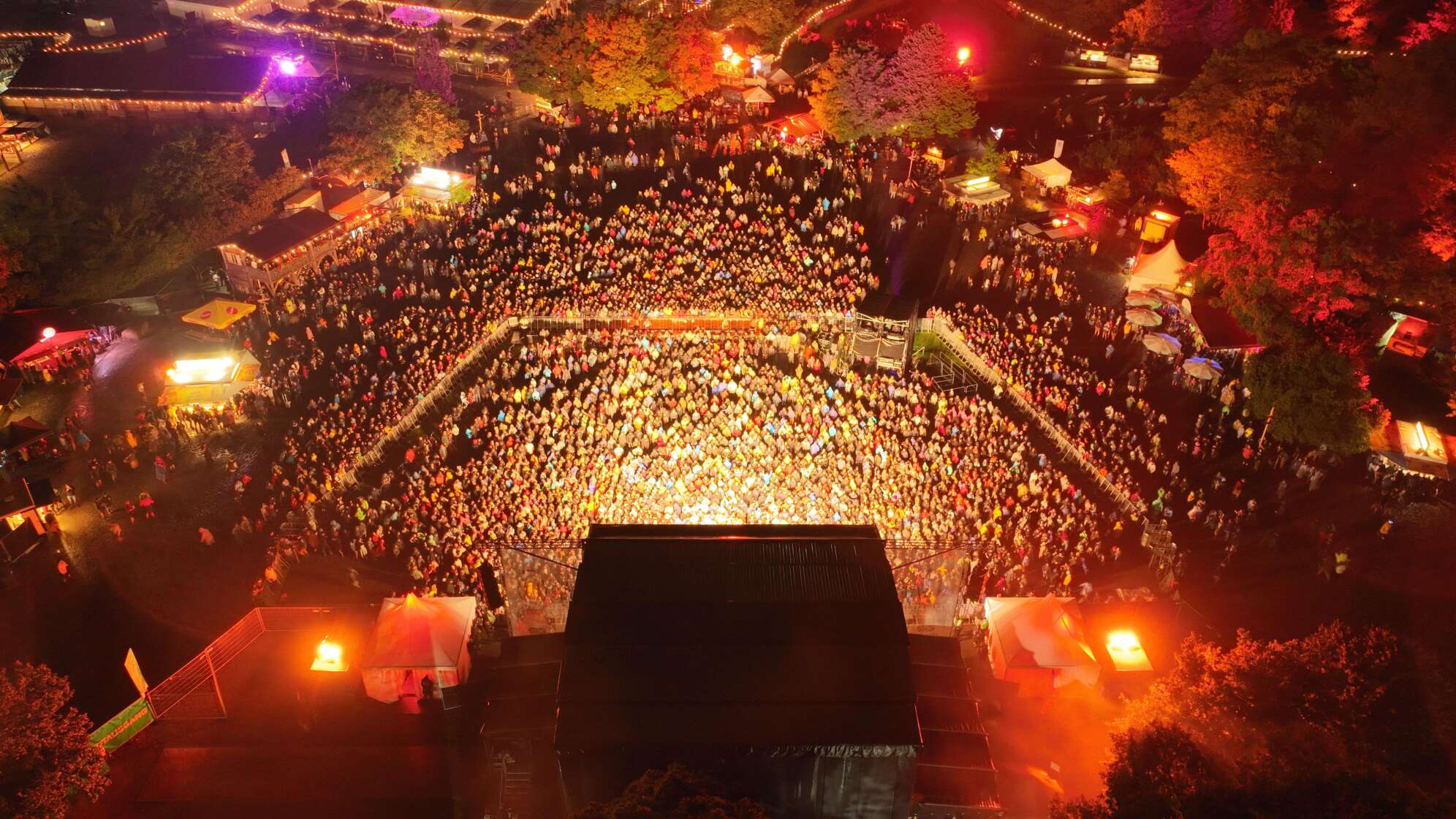 Luftaufnahme der Brass Wiesn bei Nacht. Vor einer hell beleuchteten Bühne versammelt sich eine dichte Menschenmenge, umgeben von Bäumen, Ständen und warmen Lichtern.