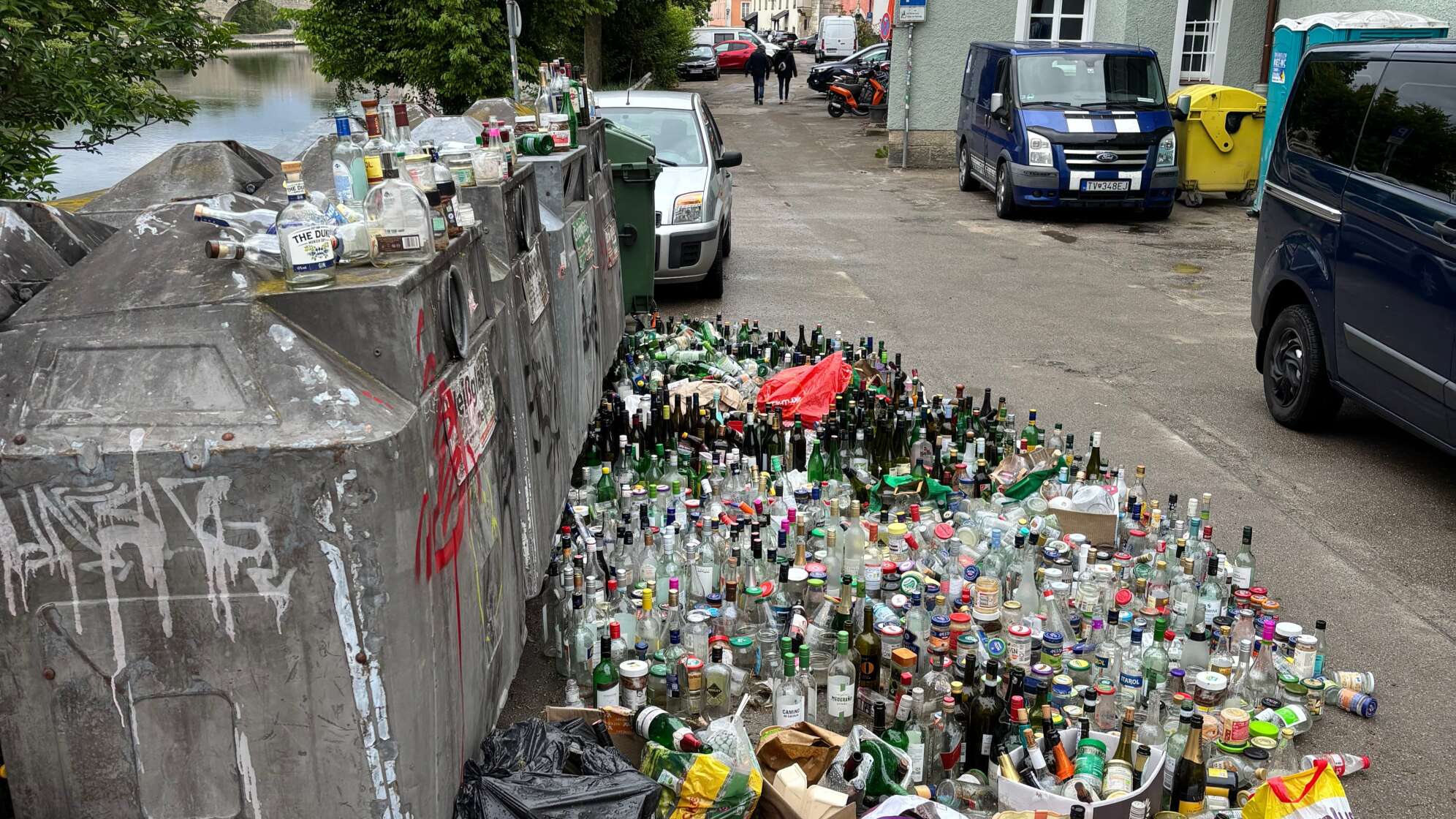 Überfüllter Müllcontainer an einem Flussufer in einer Stadt. Hunderte leere Glasflaschen, Kartons und Müllsäcke liegen neben überquellenden Containern auf der Straße, während im Hintergrund Wohnhäuser und parkende Fahrzeuge zu sehen sind.