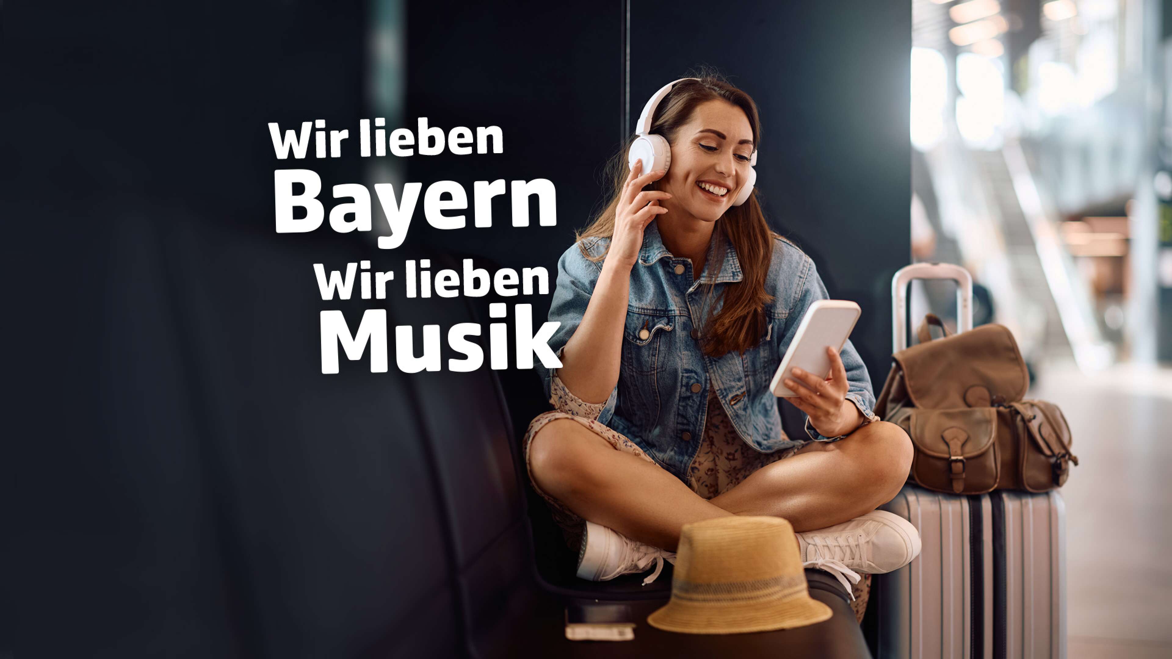 Junge Frau sitzt entspannt mit Kopfhörern und Smartphone an einem Bahnhof oder Flughafen in Bayern. Neben ihr stehen ein Koffer, ein Rucksack und ein Strohhut. Auf dem Bild steht der Text: ‚Wir lieben Bayern, wir lieben Musik‘. Moderne Mobilität, Musikgenuss und Lebensfreude in Bayern.