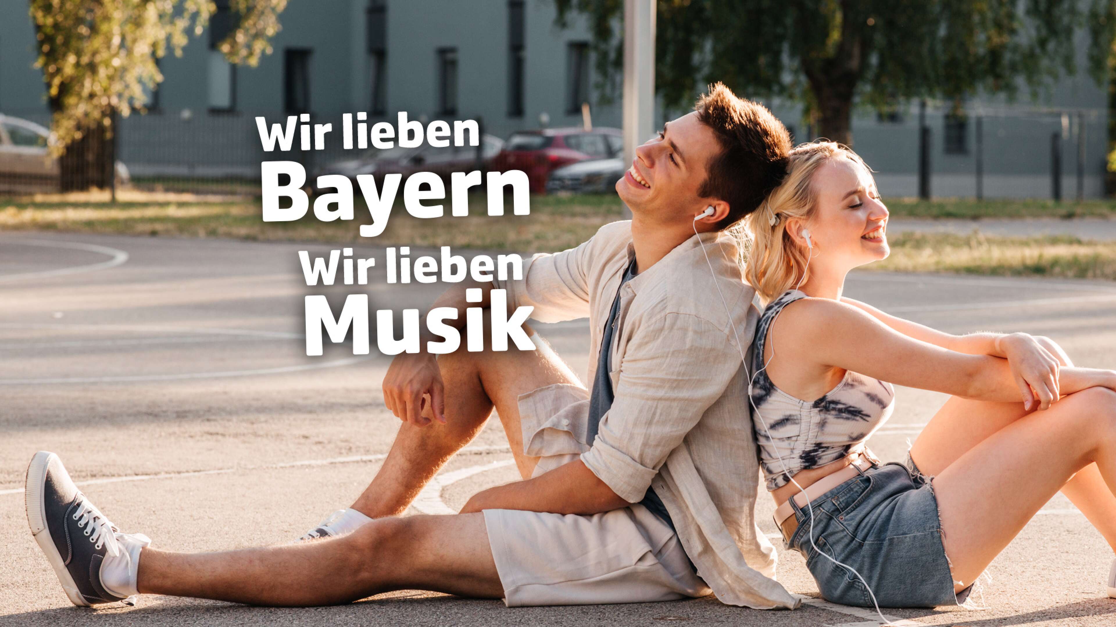Junges Paar sitzt Rücken an Rücken auf einem Basketballplatz, beide hören gemeinsam Musik mit Kopfhörern. Im Bild steht der Text: ‚Wir lieben Bayern, wir lieben Musik‘. Entspannte Freizeit, Sportplatz und Musikgenuss in Bayern.