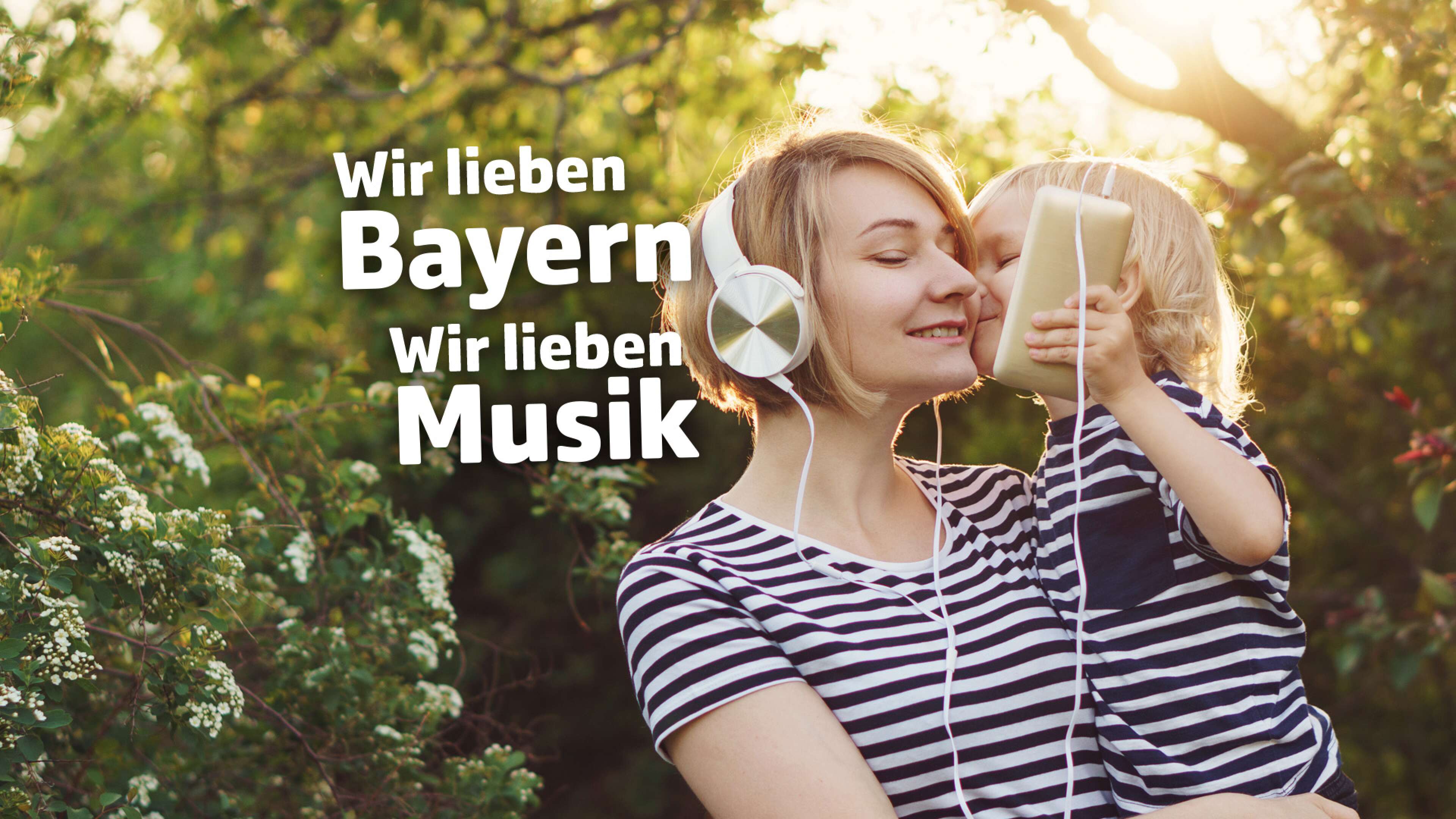 Mutter und Kind im Partnerlook mit gestreiften T-Shirts sitzen im Grünen, beide tragen Kopfhörer und hören gemeinsam Musik über ein Smartphone. Im Bild steht der Text: ‚Wir lieben Bayern, wir lieben Musik‘. Familienzeit, Natur und Musikgenuss in Bayern.