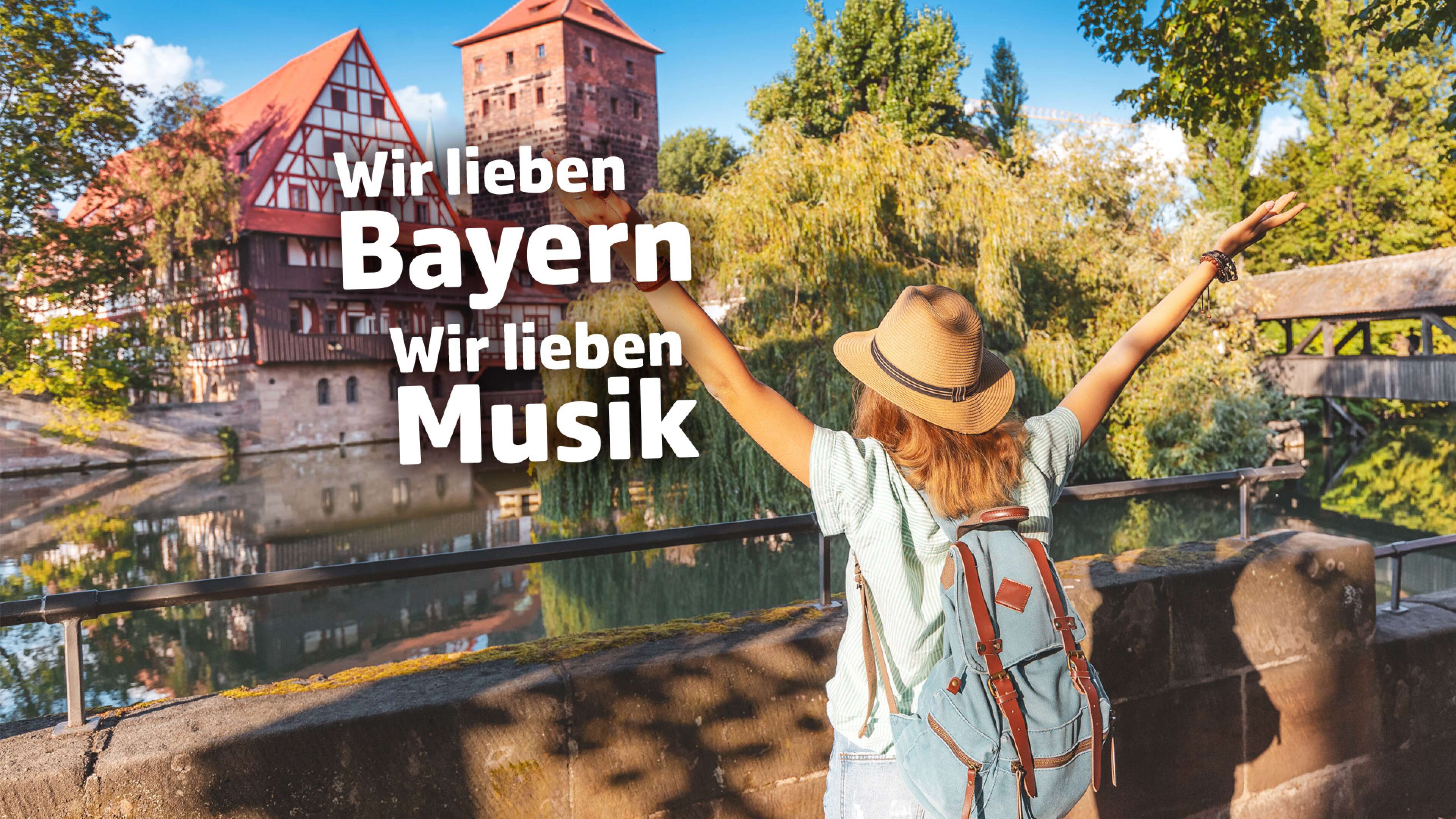 Junge Frau mit Hut und Rucksack steht mit ausgebreiteten Armen vor dem historischen Weinstadel in Nürnberg, Bayern. Im Vordergrund steht der Text: ‚Wir lieben Bayern, wir lieben Musik‘. Städtereise, Lebensfreude und Musikgenuss in Franken.