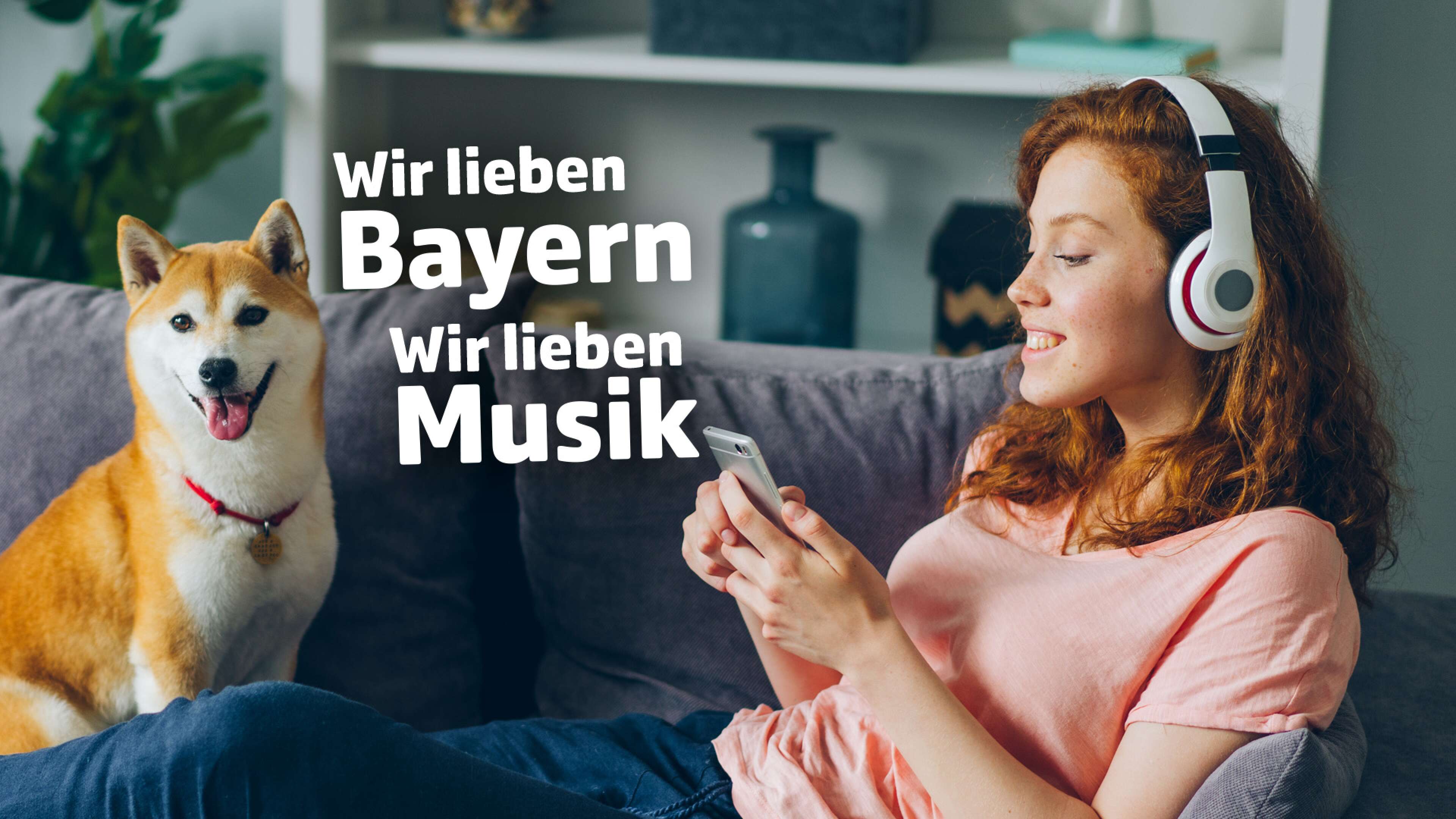 Junge Frau mit Kopfhörern liegt entspannt auf dem Sofa und hört Musik, neben ihr sitzt ein Hund. Im Bild steht der Text: ‚Wir lieben Bayern, wir lieben Musik‘. Gemütlichkeit, Haustiere und Musikgenuss zu Hause in Bayern.