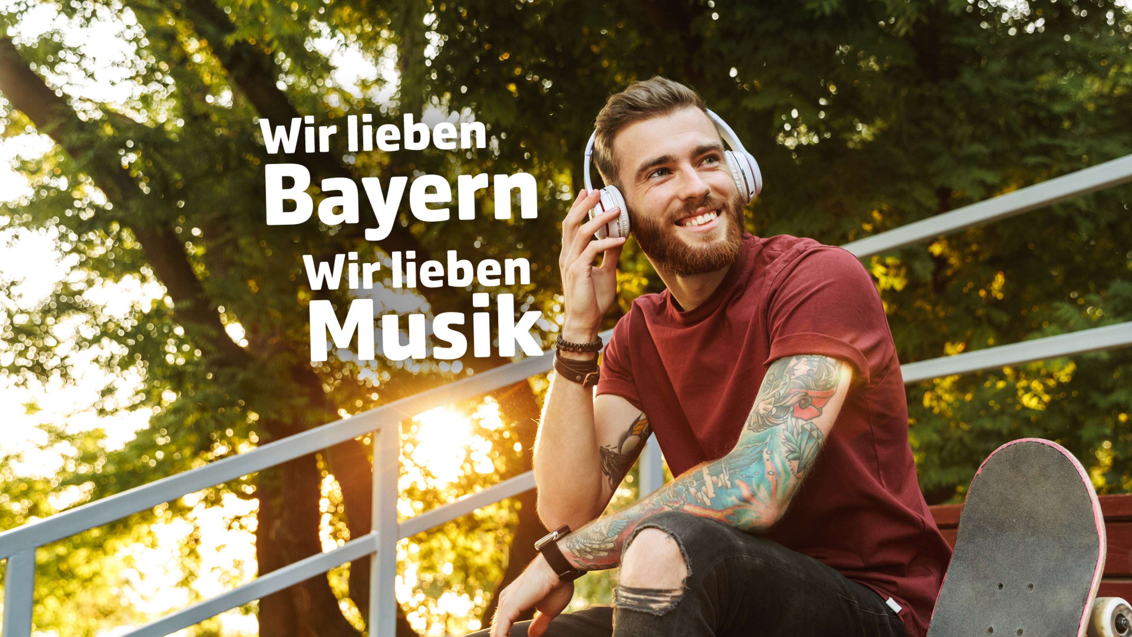 Junger Mann mit Kopfhörern und tätowiertem Arm sitzt mit Skateboard auf einer Tribüne im Park, hört Musik und genießt die Sonne. Im Bild steht der Text: ‚Wir lieben Bayern, wir lieben Musik‘. Jugendkultur, Freizeit und Musikgenuss in Bayern.