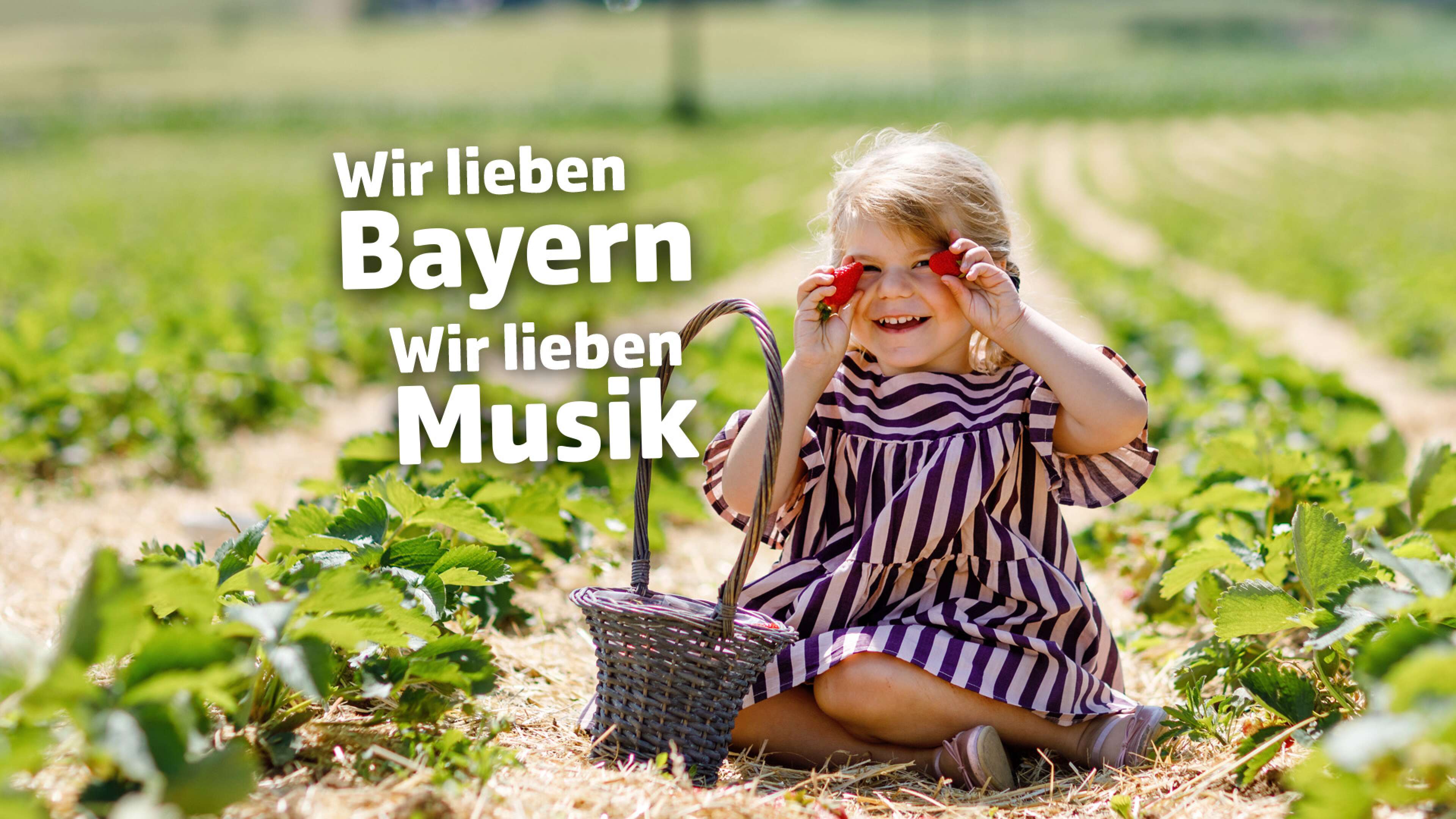 Kleines Mädchen im gestreiften Kleid sitzt auf einem Erdbeerfeld in Bayern, isst Erdbeeren und hält einen Korb. Im Bild steht der Text: ‚Wir lieben Bayern, wir lieben Musik‘. Landleben, Sommer und Musikgenuss in Bayern.