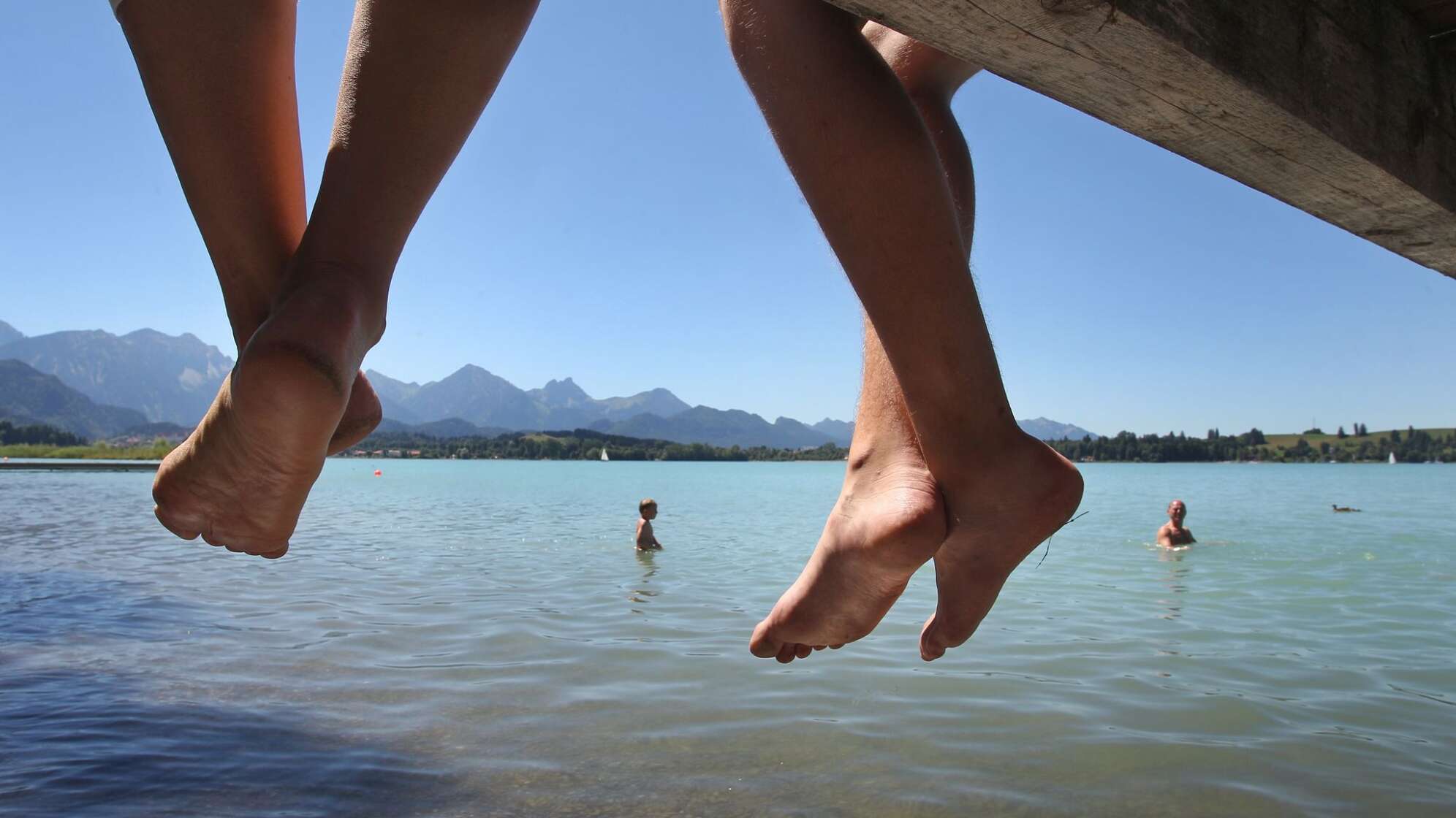 Badegäste lassen auf einem Steg am Forggensee Ihre Beine baumeln
