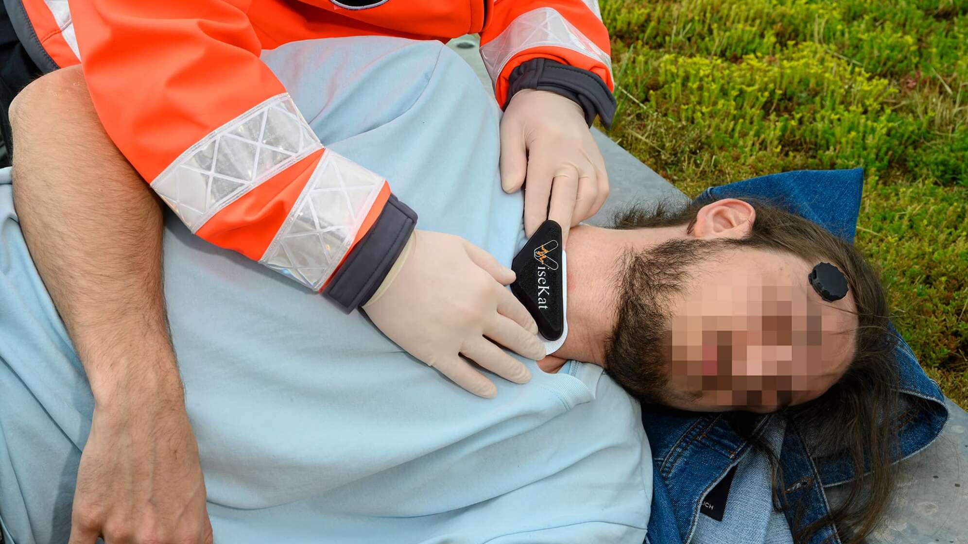 Symbolfoto zum Forschungsprojekt ViseKat des Fraunhofer IIS: Rettungskraft in orangefarbener Einsatzjacke legt einem bewusstlosen Patienten, dessen Gesicht verpixelt ist, ein sensorbasiertes Messgerät am Hals an, weiteres Sensorpflaster an der Schläfe – Entwicklung digitaler Systeme zur Vitaldaten-Erfassung und Überwachung von Patienten in der Notfallmedizin.