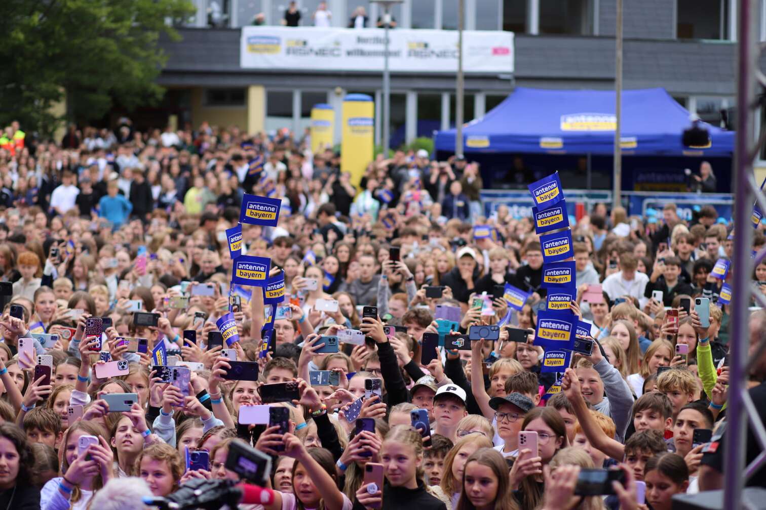 Popstar LOI am 23.07.2026 live auf der Bühne beim zweiten ANTENNE BAYERN Schulhof-Gig 2026 auf dem Pausenhof der Staatlichen Realschule in Neustadt bei Coburg in Oberfranken - Foto vom Konzert mit fröhlichen Schülern, hochgehaltenen Smartphones und Plakaten