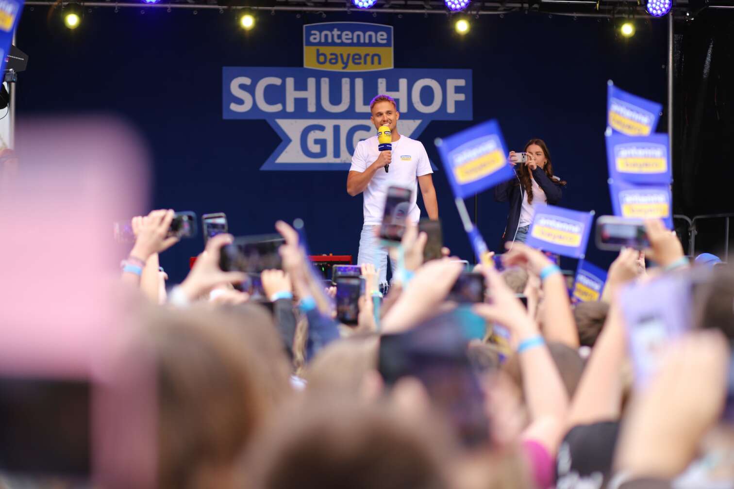 Popstar LOI am 23.07.2026 live auf der Bühne beim zweiten ANTENNE BAYERN Schulhof-Gig 2026 auf dem Pausenhof der Staatlichen Realschule in Neustadt bei Coburg in Oberfranken - Foto vom Konzert mit fröhlichen Schülern, hochgehaltenen Smartphones und Plakaten