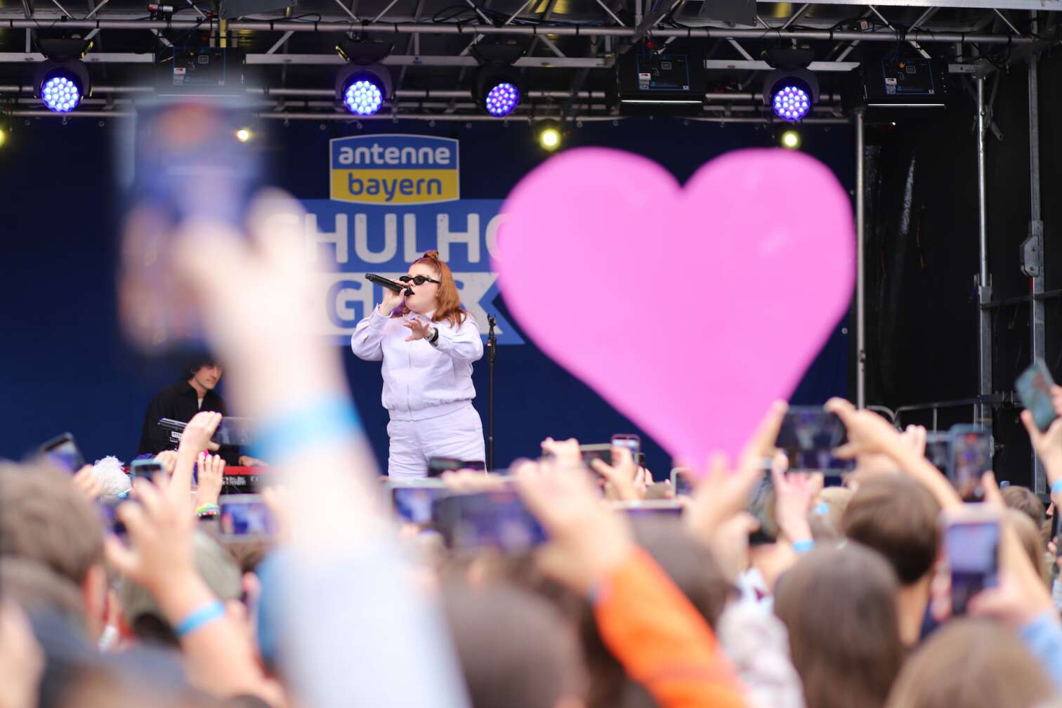Popstar LOI am 23.07.2026 live auf der Bühne beim zweiten ANTENNE BAYERN Schulhof-Gig 2026 auf dem Pausenhof der Staatlichen Realschule in Neustadt bei Coburg in Oberfranken - Foto vom Konzert mit fröhlichen Schülern, hochgehaltenen Smartphones und Plakaten