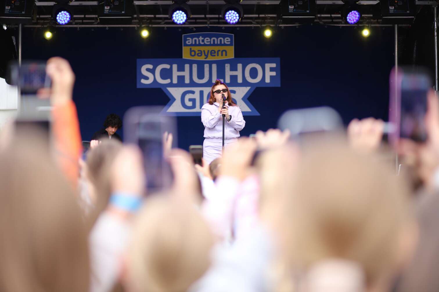 Popstar LOI am 23.07.2026 live auf der Bühne beim zweiten ANTENNE BAYERN Schulhof-Gig 2026 auf dem Pausenhof der Staatlichen Realschule in Neustadt bei Coburg in Oberfranken - Foto vom Konzert mit fröhlichen Schülern, hochgehaltenen Smartphones und Plakaten