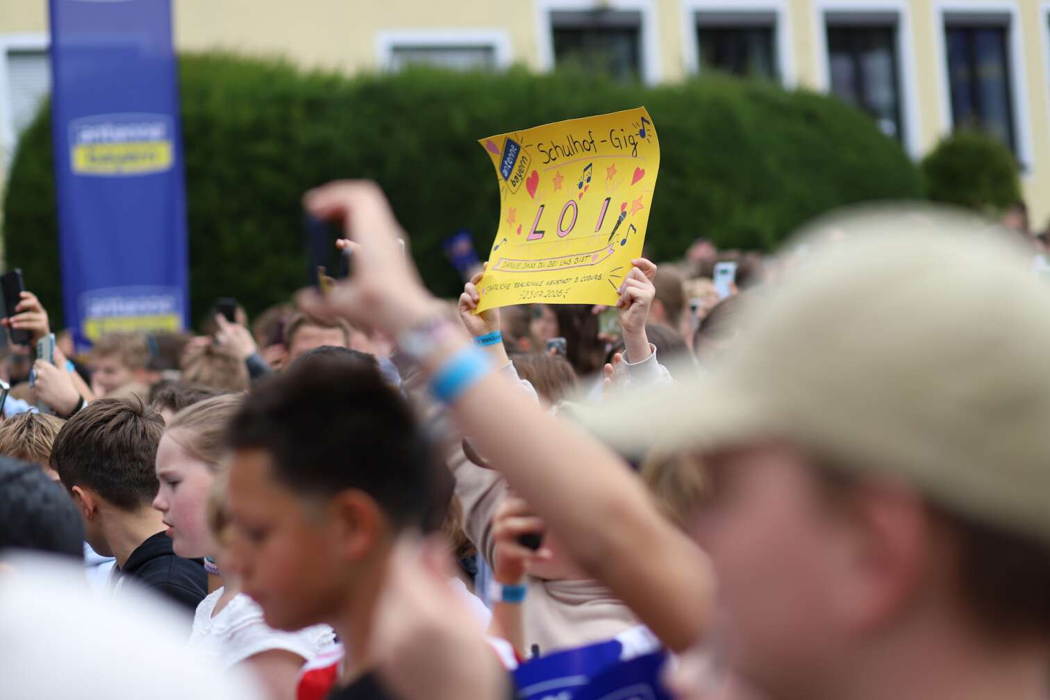 Popstar LOI am 23.07.2026 live auf der Bühne beim zweiten ANTENNE BAYERN Schulhof-Gig 2026 auf dem Pausenhof der Staatlichen Realschule in Neustadt bei Coburg in Oberfranken - Foto vom Konzert mit fröhlichen Schülern, hochgehaltenen Smartphones und Plakaten