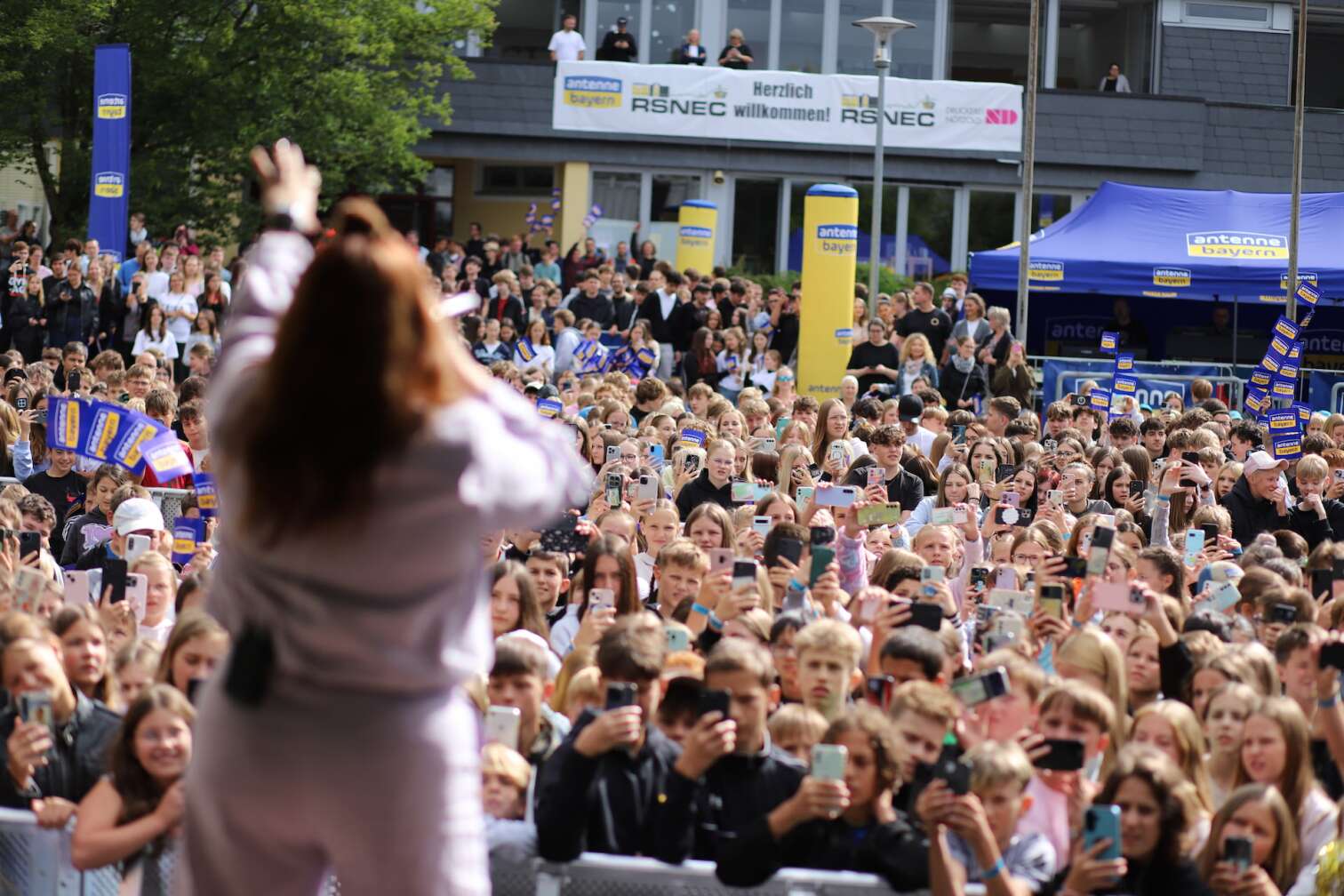 Popstar LOI am 23.07.2026 live auf der Bühne beim zweiten ANTENNE BAYERN Schulhof-Gig 2026 auf dem Pausenhof der Staatlichen Realschule in Neustadt bei Coburg in Oberfranken - Foto vom Konzert mit fröhlichen Schülern, hochgehaltenen Smartphones und Plakaten