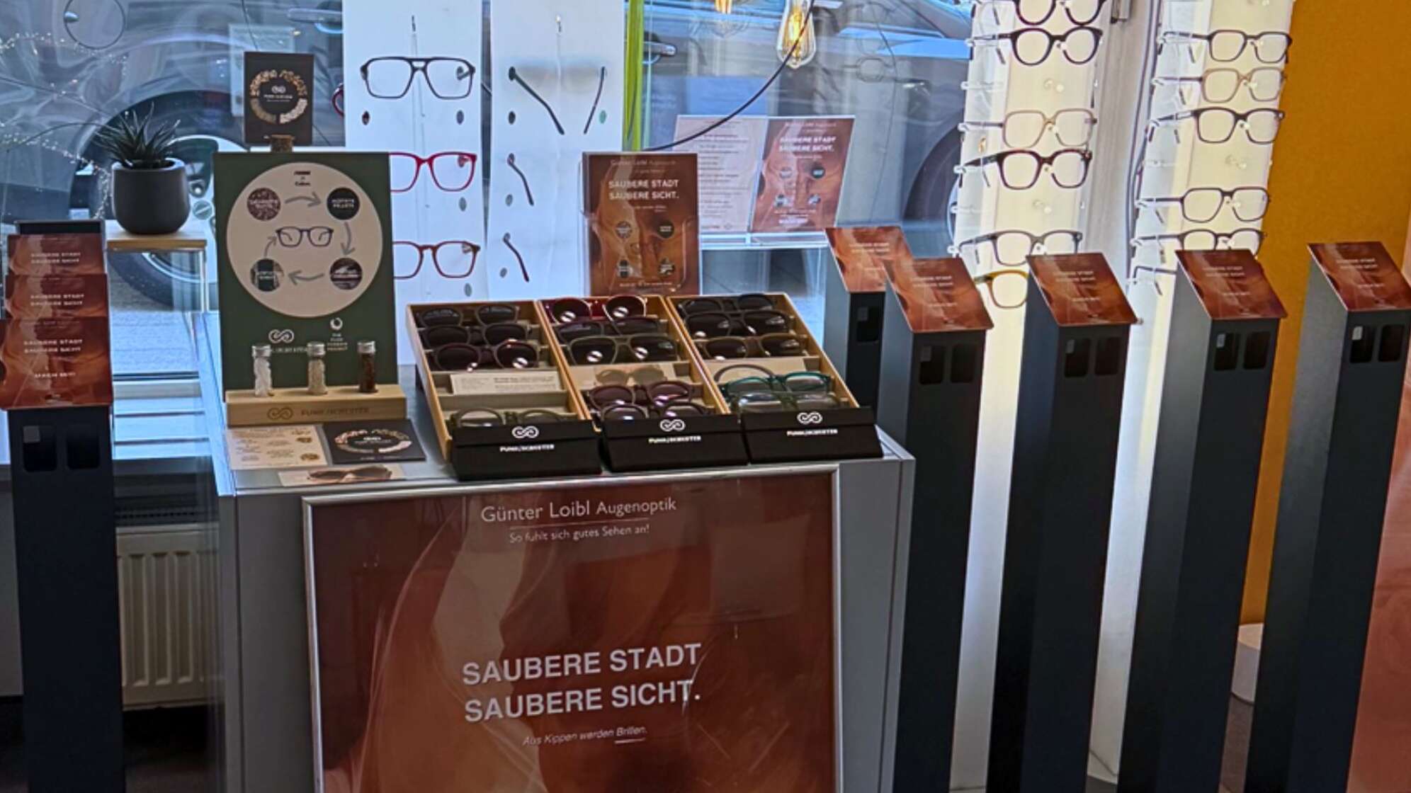 Schaufenster von „Günter Loibl Augenoptik“ in Pocking mit nachhaltiger Brillenkollektion: mehrere Tabletts mit modernen Brillenfassungen, Infotafeln und Gläser mit recycelten Materialien, Slogan „Saubere Stadt, saubere Sicht – aus Kippen werden Brillen“. Präsentation eines Umweltprojekts zur Brillenherstellung aus Zigarettenabfällen in Pocking, Bayern.