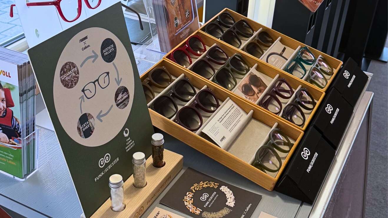 Schaufenster von „Günter Loibl Augenoptik“ in Pocking mit nachhaltiger Brillenkollektion: mehrere Tabletts mit modernen Brillenfassungen, Infotafeln und Gläser mit recycelten Materialien, Slogan „Saubere Stadt, saubere Sicht – aus Kippen werden Brillen“. Präsentation eines Umweltprojekts zur Brillenherstellung aus Zigarettenabfällen in Pocking, Bayern.