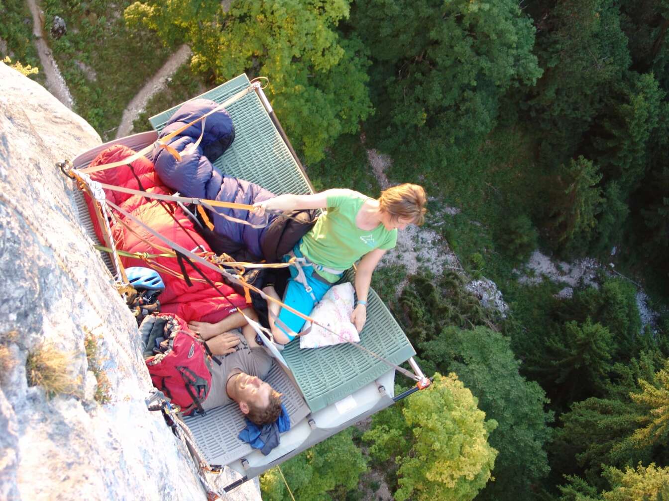 Extremes Übernachtungs-Erlebnis in Bayern: Frau und Mann liegen mit Ausrüstung und Schlafsäcken auf einer gesicherten Metallplattform direkt an einer Felswand, tief unter ihnen dichter Wald in den Alpen – Steilwand-Schlafen für Mutige.