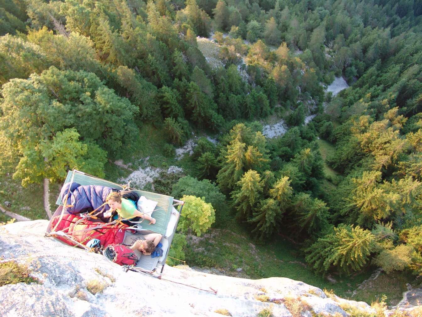 Luftaufnahme Steilwand-Schlafen in den Bayerischen Alpen: Outdoor-Schlafplattform hängt an der Felswand hoch über einem grünen Bergwald, zwei Abenteurer mit Schlafsäcken genießen den Ausblick – spektakuläre Übernachtung am Fels in Bayern.