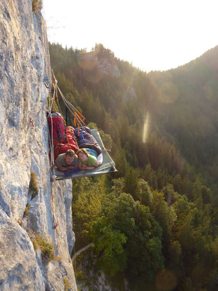 Steilwand-Schlafen in Bayern: zwei Personen liegen in einer Metallplattform mit Schlafsäcken an einer senkrechten Felswand über einem dichten Bergwald, Sonnenuntergang und Fernblick auf die Alpen – außergewöhnliches Übernachtungs-Abenteuer für Kletterer.