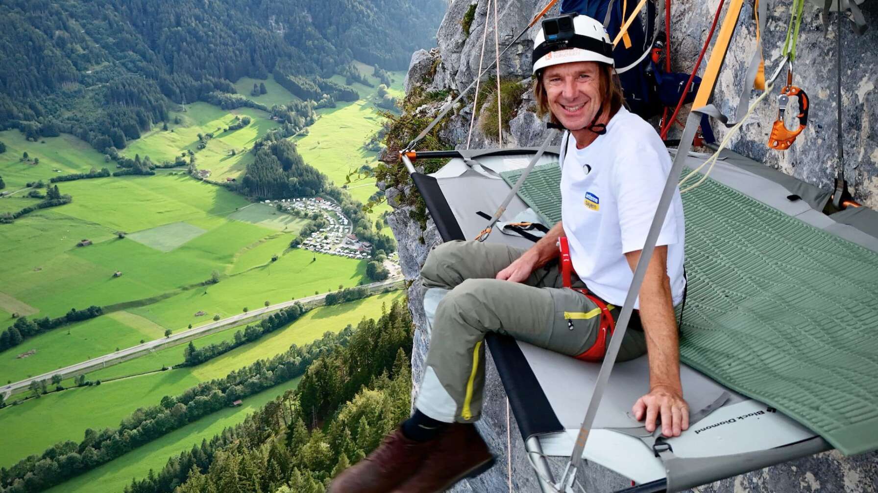 Wolfgang Leikermoser sitzt auf einer Portaledge Hängematte, die an einer Felswand in den Allgäuer Alpen hängt und lächelt Richtung Kamera