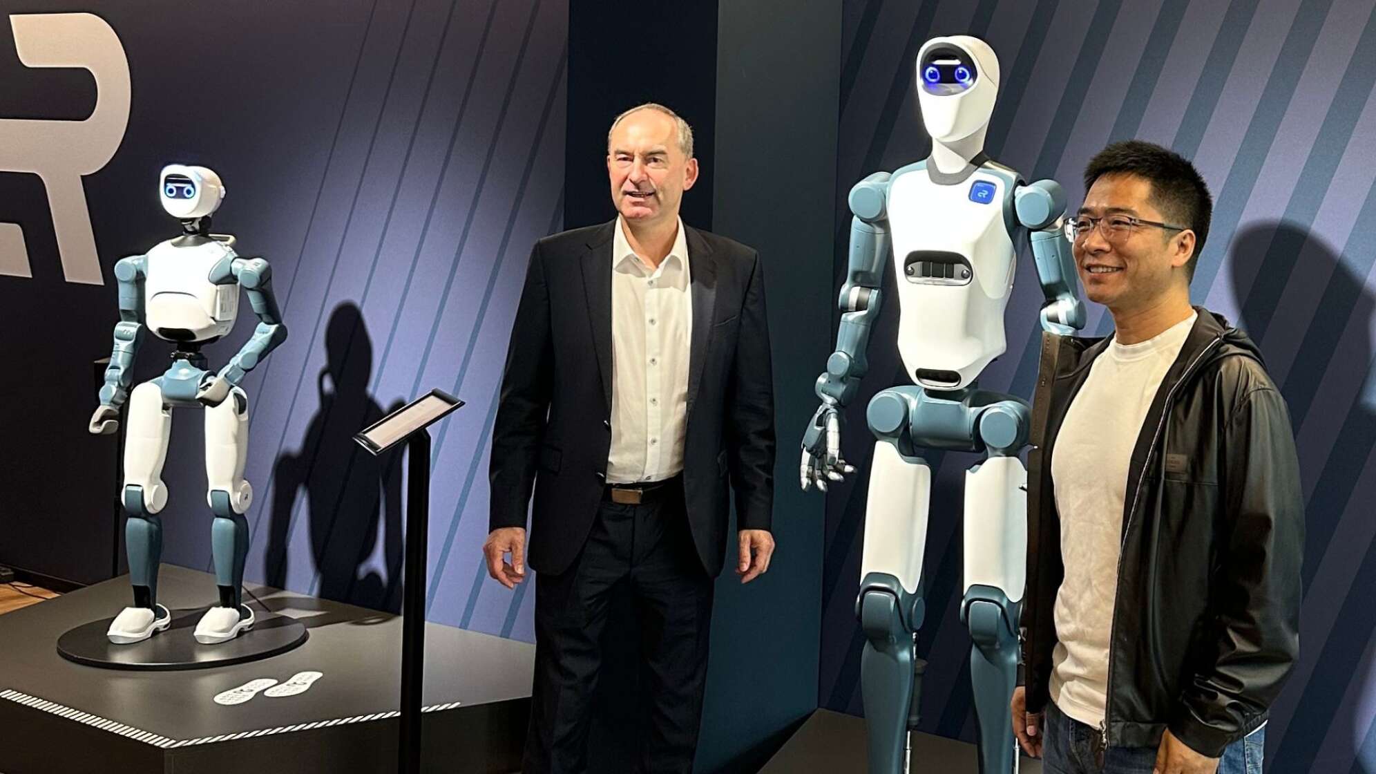 Besuch bei Agile Robots in Bayern: zwei Männer stehen zwischen zwei humanoiden Industrierobotern in Ausstellungsraum, moderne weiß-blaue Robotertechnik mit Kamerakopf und Greifhänden, Symbolbild für KI, Robotik und Zukunft der Industrie in Deutschland.