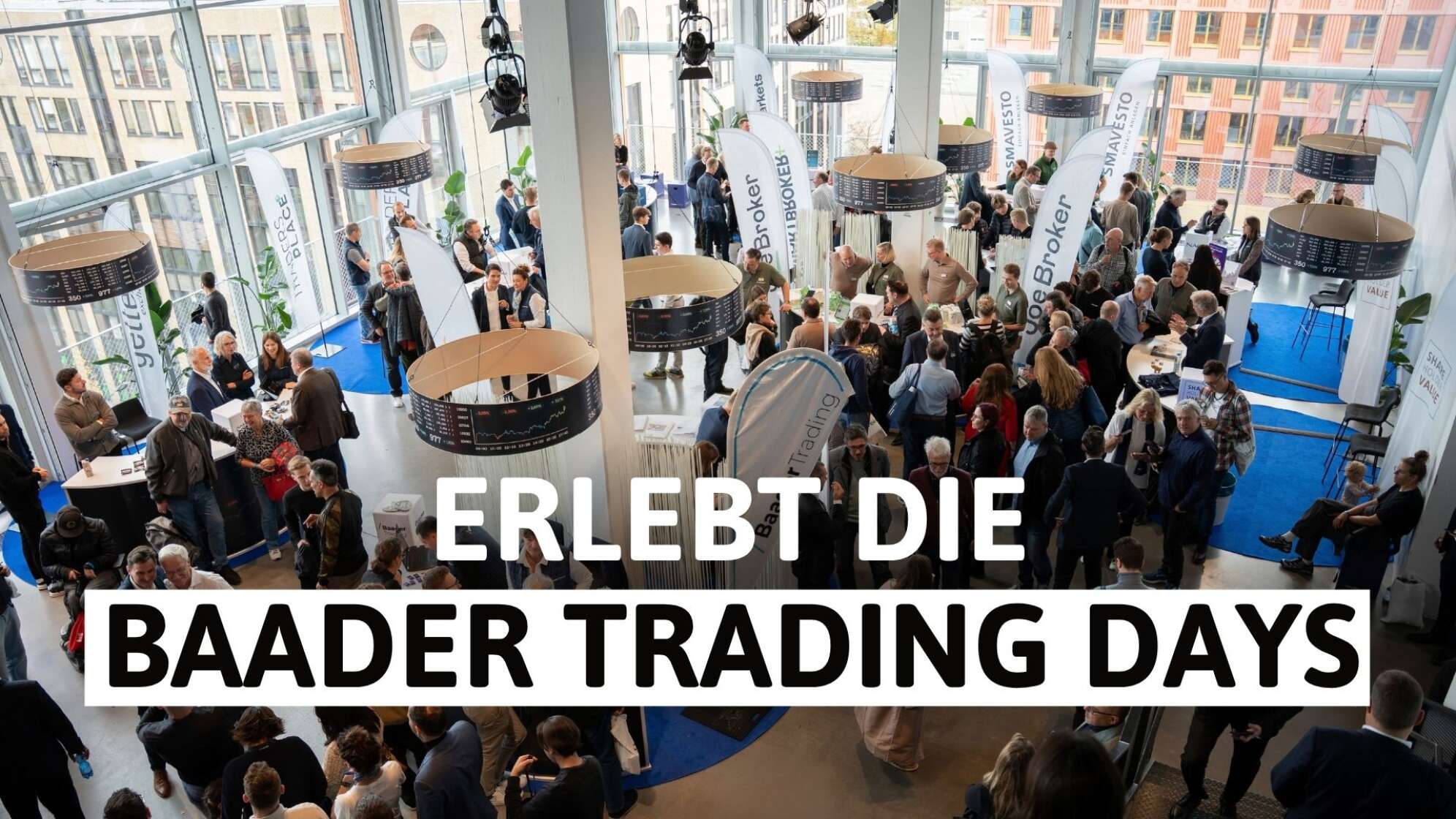 Veranstaltungsfoto von den Baader Trading Days 2026: viele Besucherinnen und Besucher im modernen Messe- und Foyerbereich mit großen Fensterfronten, Infoständen und Trading-Partner-Flaggen, darüber große weiße Schrift „Erlebt die Baader Trading Days“ – Börsen- und Finanzmesse in Deutschland.