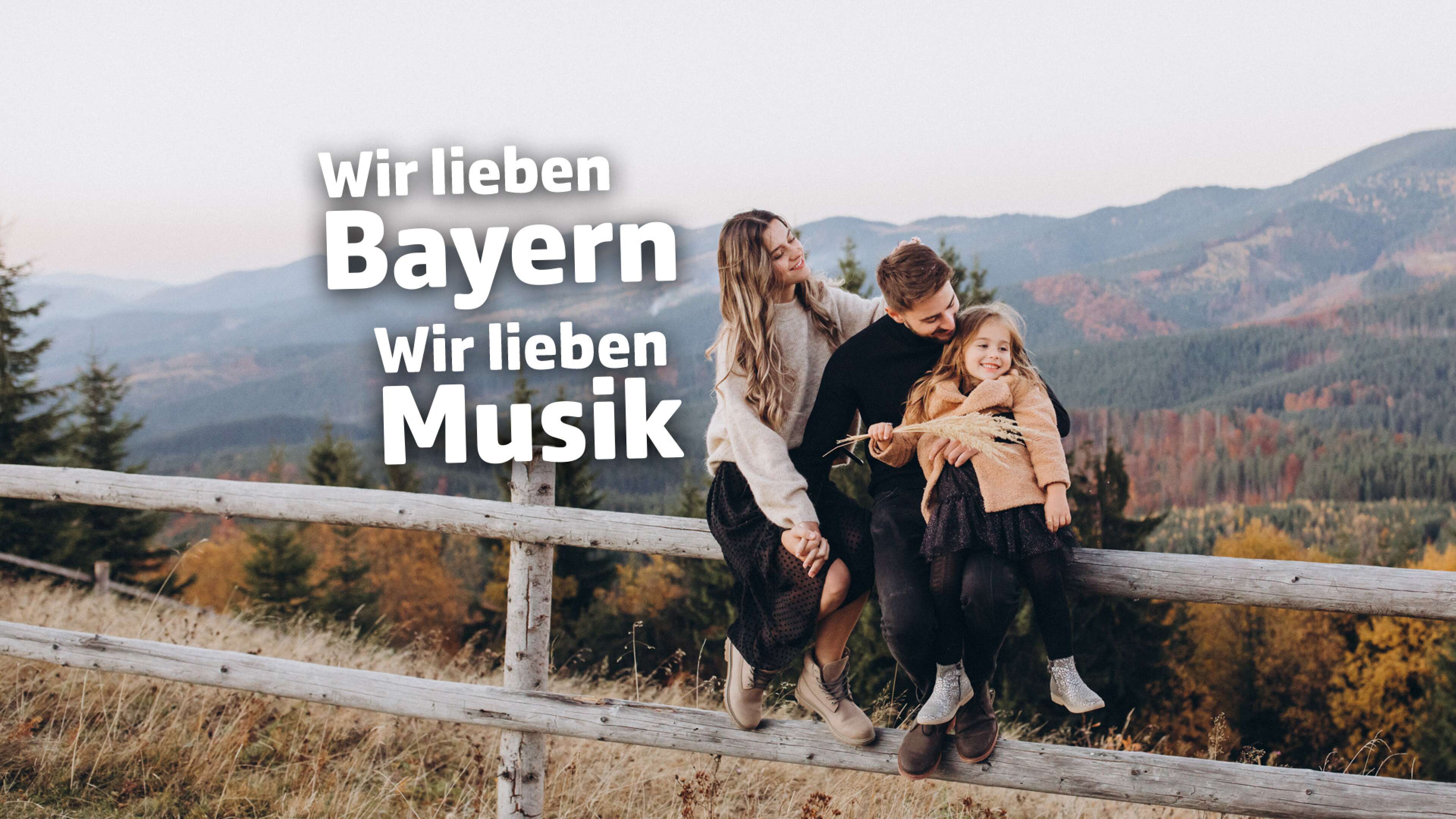 Junge Familie sitzt lachend auf einem Holzzaun mit Blick auf herbstliche Berge in Bayern; links eingeblendet der Text ‚Wir lieben Bayern – Wir lieben Musik‘ als Imagewerbung für den Radiosender ANTENNE BAYERN.