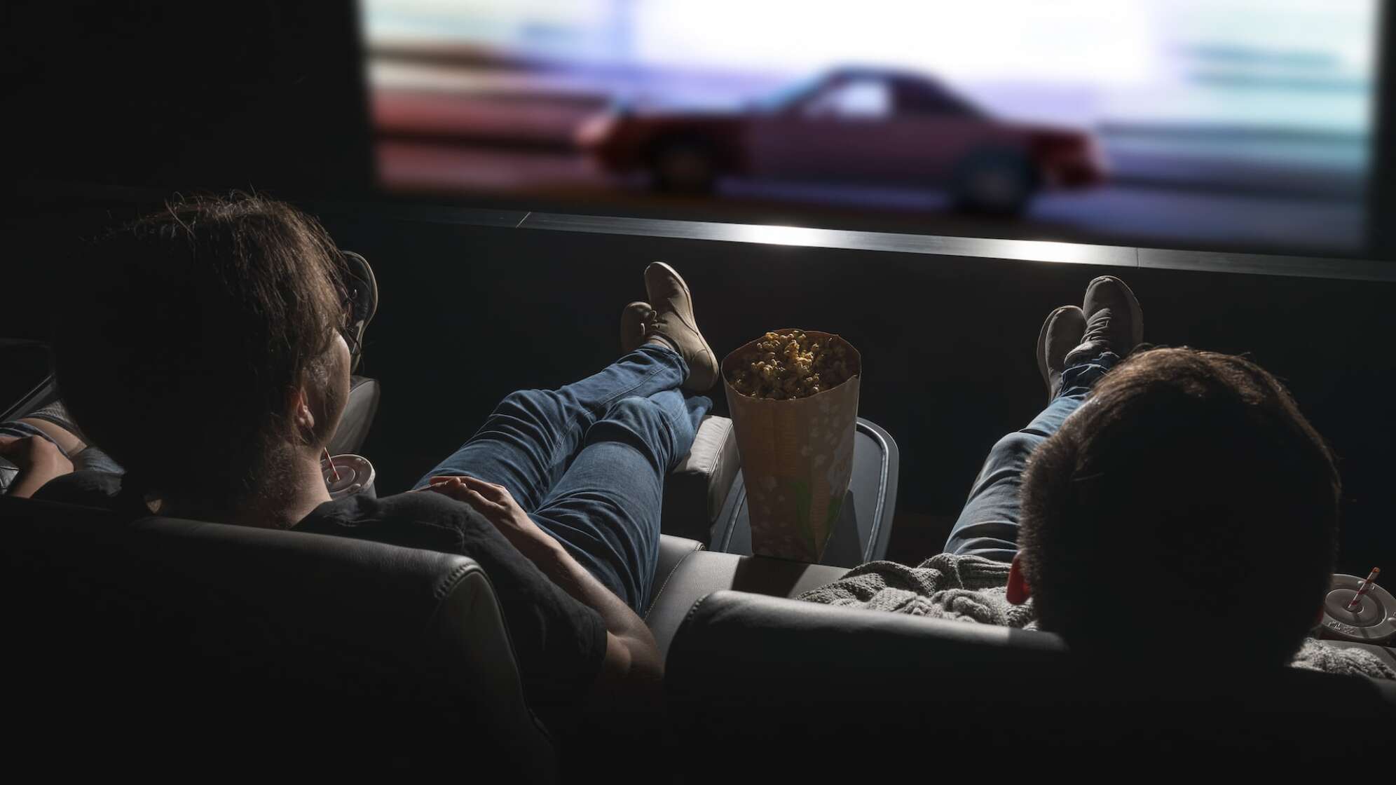 Foto von zwei Kinobesuchern von Hinten in entspannter Position, zwischen sich Popcorn und Getränke