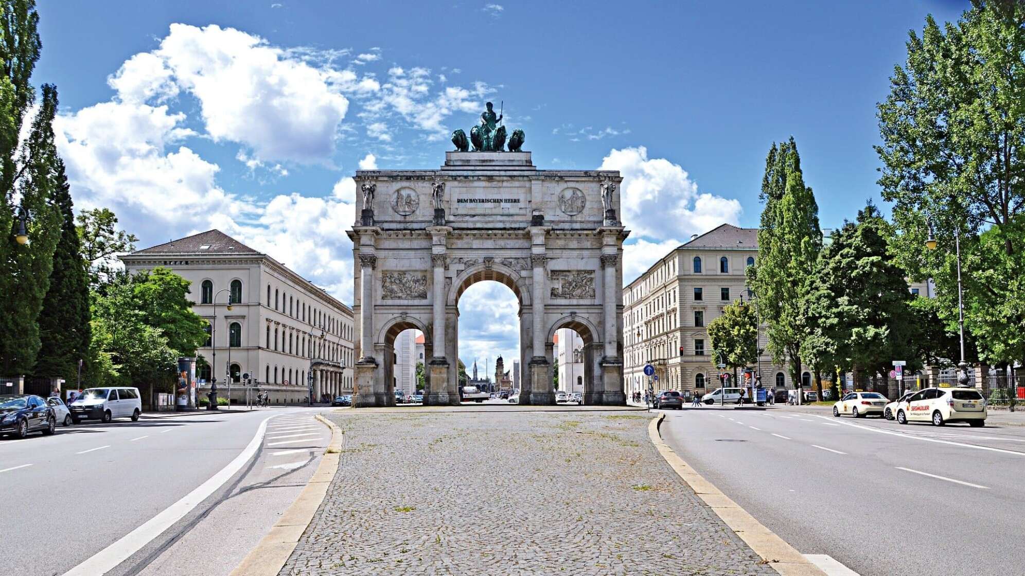 Siegestor in München bei klarem Himmel, umgeben von historischen Gebäuden und Bäumen