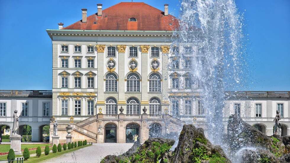 Schloss Nymphenburg in München mit reflektierendem Wasser im Vordergrund und blauem Himmel