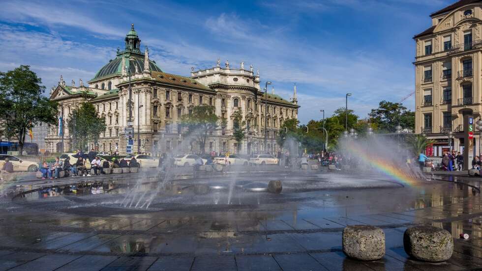 Karlsplatz in München mit Brunnen, historischen Gebäuden und Menschen bei sonnigem Wetter.